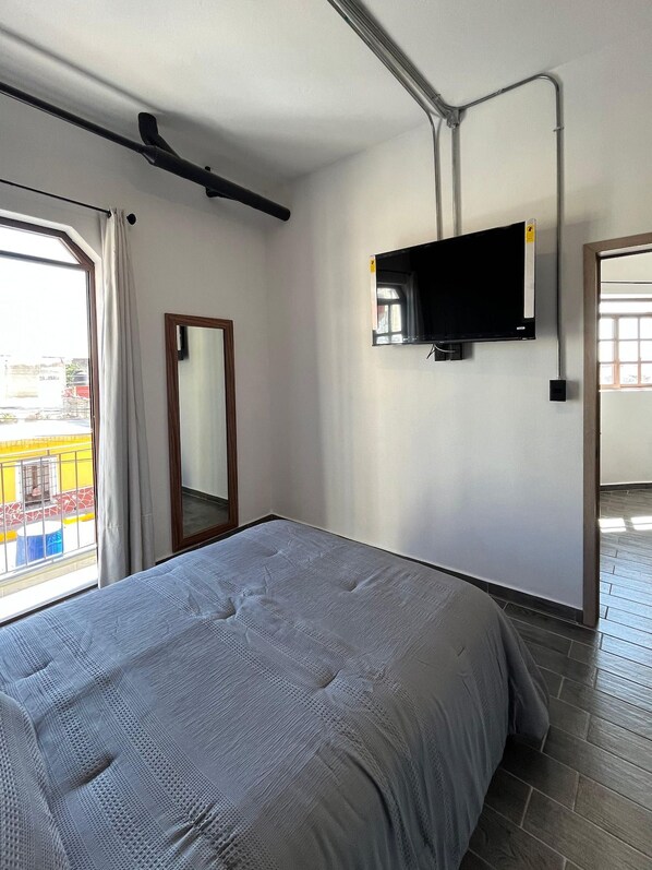 A bedroom with a bed, a flat-screen TV mounted on the wall, a window with curtains, and a view of the outside.