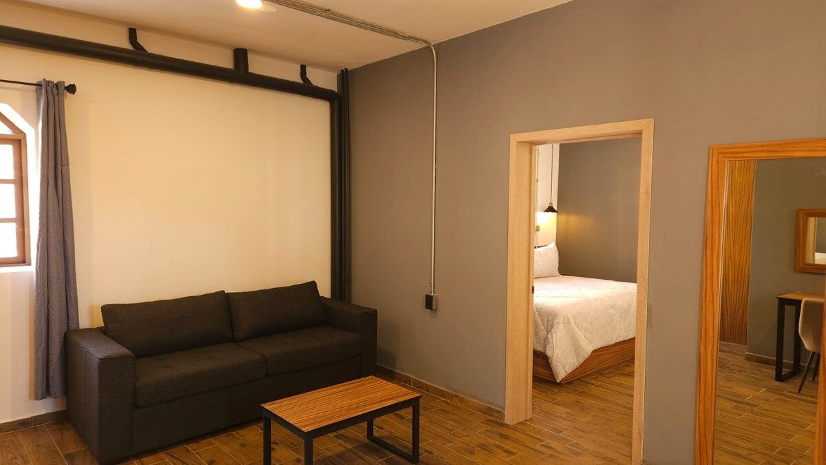 Quarto com sofá de cor escura, uma pequena mesa de madeira, cama com colcha branca e piso de madeira.