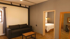 A room with a dark-colored sofa, a small wooden table, a bed with a white bedspread, and a wooden floor.