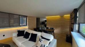 A modern living room with a sofa, a dining table, and chairs. There is a window with blinds, a kitchen area in the background, and a large mirror on the wall. - Sixth floor facing Parque España, Condesa (Mexico City)