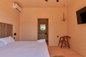 A bedroom with a bed, a wooden headboard, a small wooden stool, a wall-mounted TV, and an air conditioning unit. - Ocean View Suite w/ Rooftop & Pool Access (Puerto Escondido)