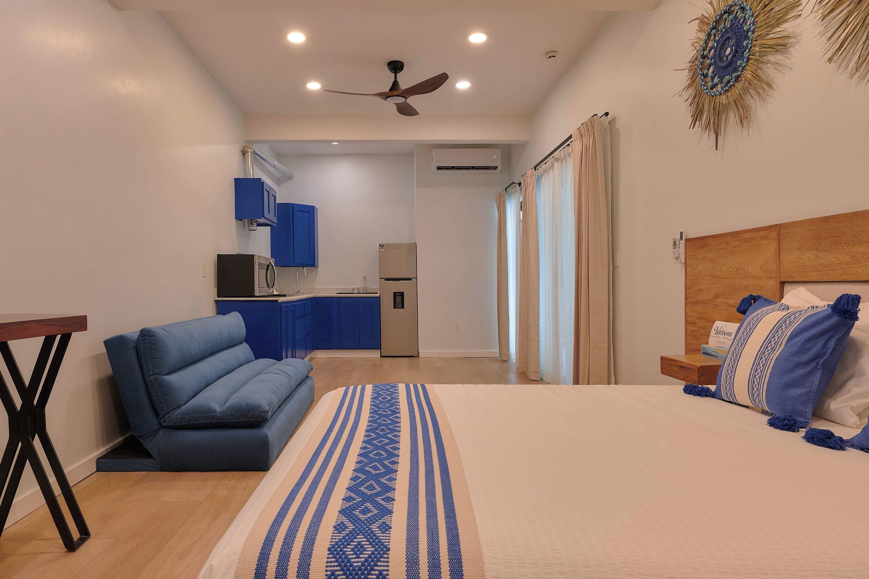 A bedroom with a bed, a sofa, a small table, a ceiling fan, and a kitchenette in the background.