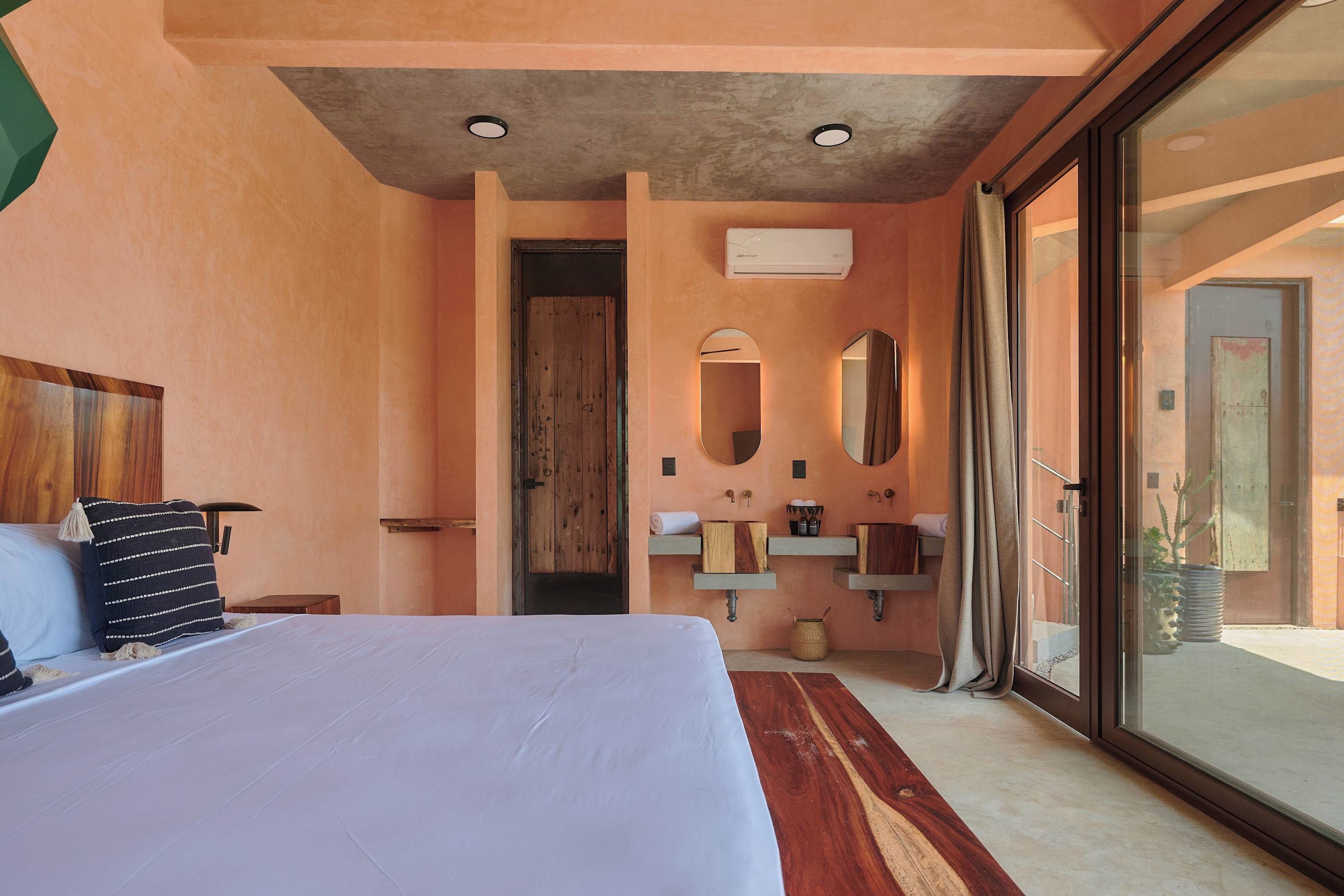Ein Schlafzimmer mit einem großen Bett, einem Holz-Kopfstück, einem Nachttisch, einem Fenster mit Blick und einem Badezimmer mit Waschbecken, Spiegel und Handtuchhalter.
