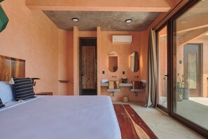 Ein Schlafzimmer mit einem großen Bett, einem Holz-Kopfstück, einem Nachttisch, einem Fenster mit Blick und einem Badezimmer mit Waschbecken, Spiegel und Handtuchhalter.