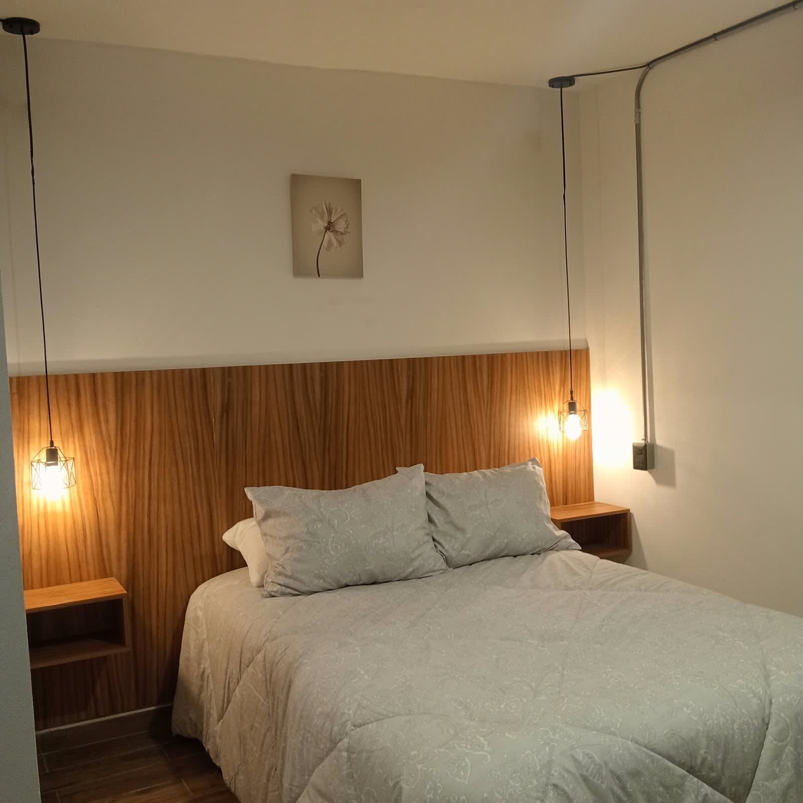 Ett sovrum med en sänggavel i trä, en säng med ett ljust täcke, två sängbord och en väggmonterad lampa.