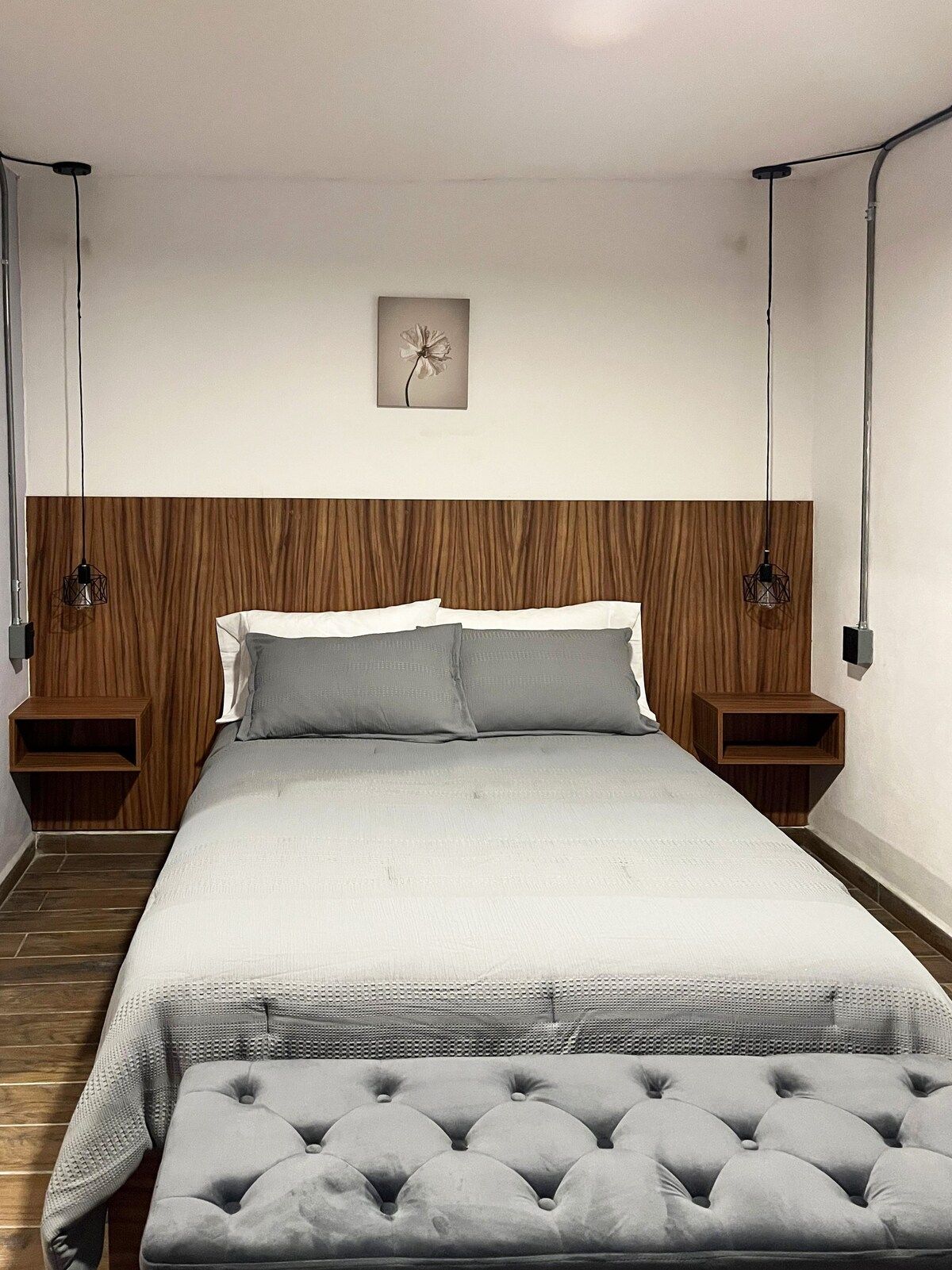 Quarto com cabeceira de madeira, cama com roupa de cama branca, duas mesas de cabeceira, luminária na parede e uma única obra de arte.