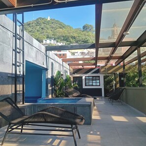 A rooftop terrace with a pool, lounge chairs, and a view of a hill with buildings.