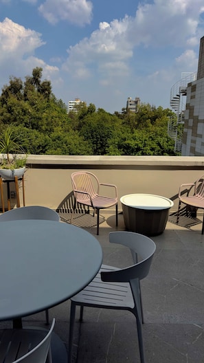 An outdoor seating area with round tables and chairs, a potted plant, and a view of a cityscape with buildings and trees. - Eighth floor facing Parque España, Condesa (Ciudad de México)