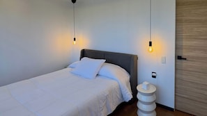 Un lit bien fait, avec une tête de lit grise, une table de chevet sur laquelle se trouve un objet blanc, et deux lampes suspendues au plafond.