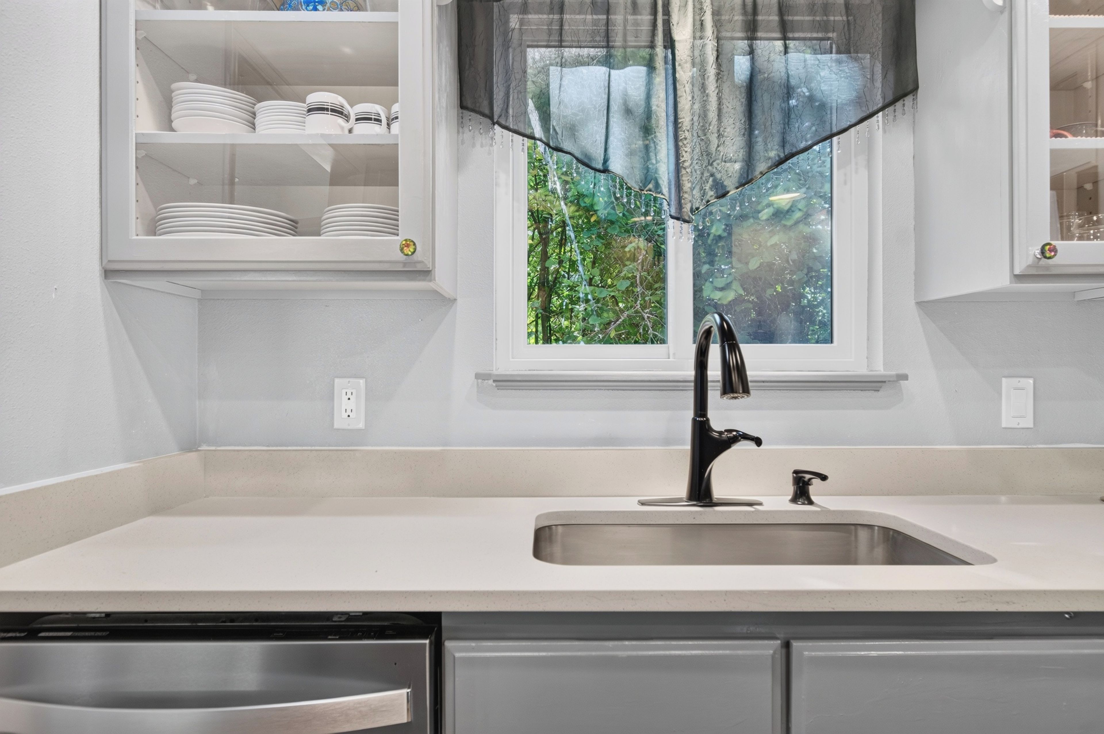 Una cocina con un lavamanos de acero inoxidable, encimeras blancas y una ventana con vistas a zonas verdes.
