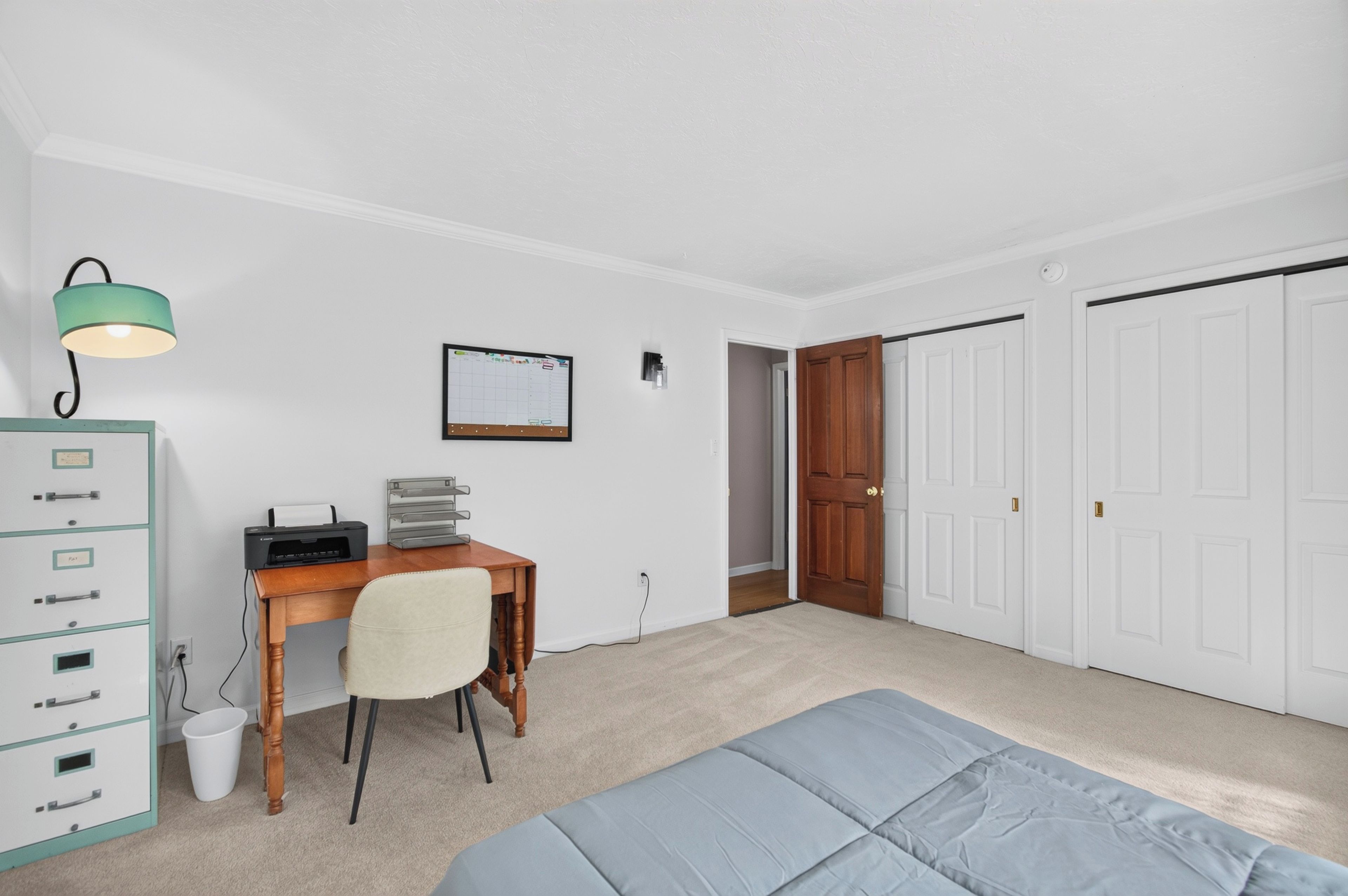 A bedroom with a desk, chair, bed, wardrobe, and a door leading to another room.