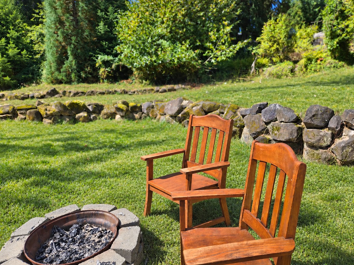 Two wooden chairs and a fire pit in a grassy outdoor area with a stone wall and trees in the background.