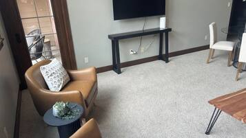 A living room with a flat-screen TV mounted on the wall, a dark wooden coffee table, and a beige carpet.