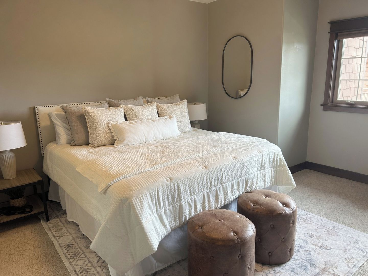 A bedroom with a large bed, two bedside lamps, a round mirror, and a window with blinds.