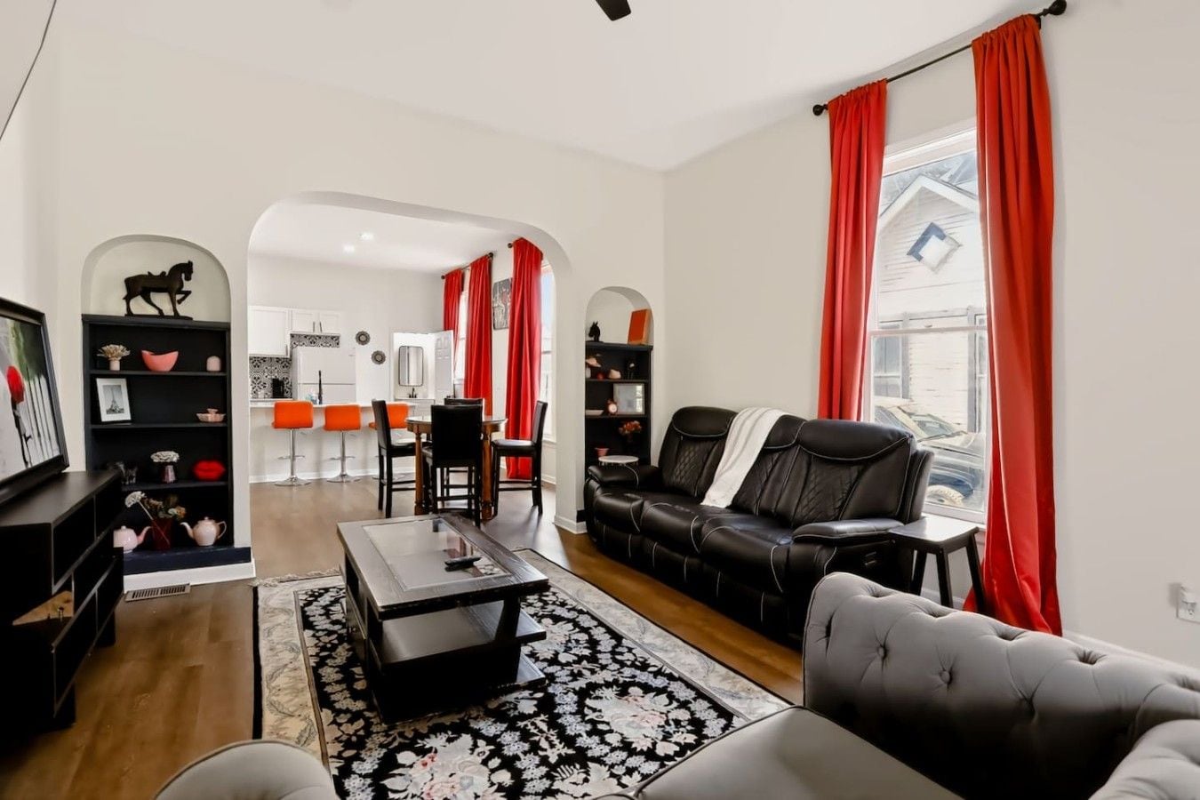 A modern living room with a black leather sofa, a coffee table, and a TV on a stand. There is a dining area with red chairs and a table, and a kitchen visible through an archway.