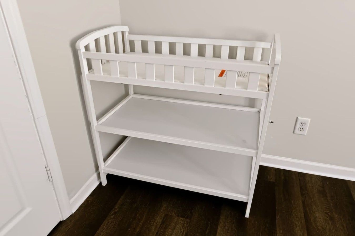A white, three-tiered, white-framed storage unit against a wall with a power outlet.