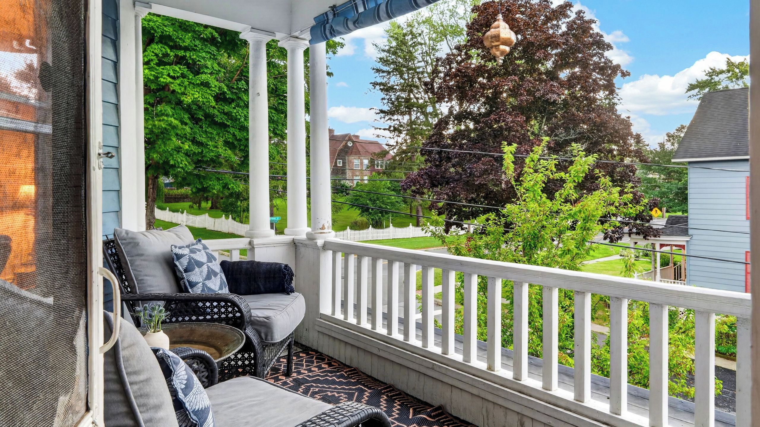 A covered porch with a white railing, a wicker sofa with cushions, and a view of a green landscape with trees and a house.