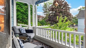 A covered porch with a white railing, a wicker sofa with cushions, and a view of a green landscape with trees and a house. - The Painted Lady by Avantstay Grand Victorian + Balcony Firepit + BBQ (Catskill)