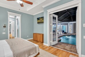 Ein Schlafzimmer mit einem Bett, einer Holzbank, einem Deckenventilator und Blick auf ein Außenbecken durch eine offene Tür.