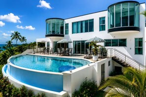 A modern two-story building with a curved swimming pool, outdoor seating, and a view of the ocean.