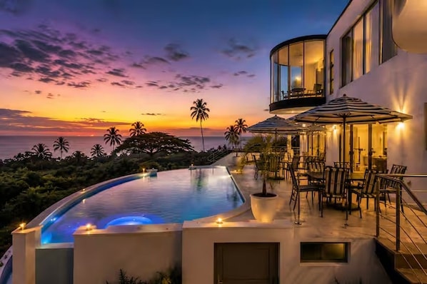 A modern building with a curved infinity pool, outdoor seating, and a view of the ocean at sunset.