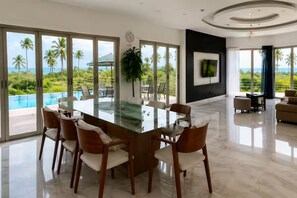 A spacious dining area with a glass table and chairs, a large potted plant, and a view of a swimming pool and palm trees.