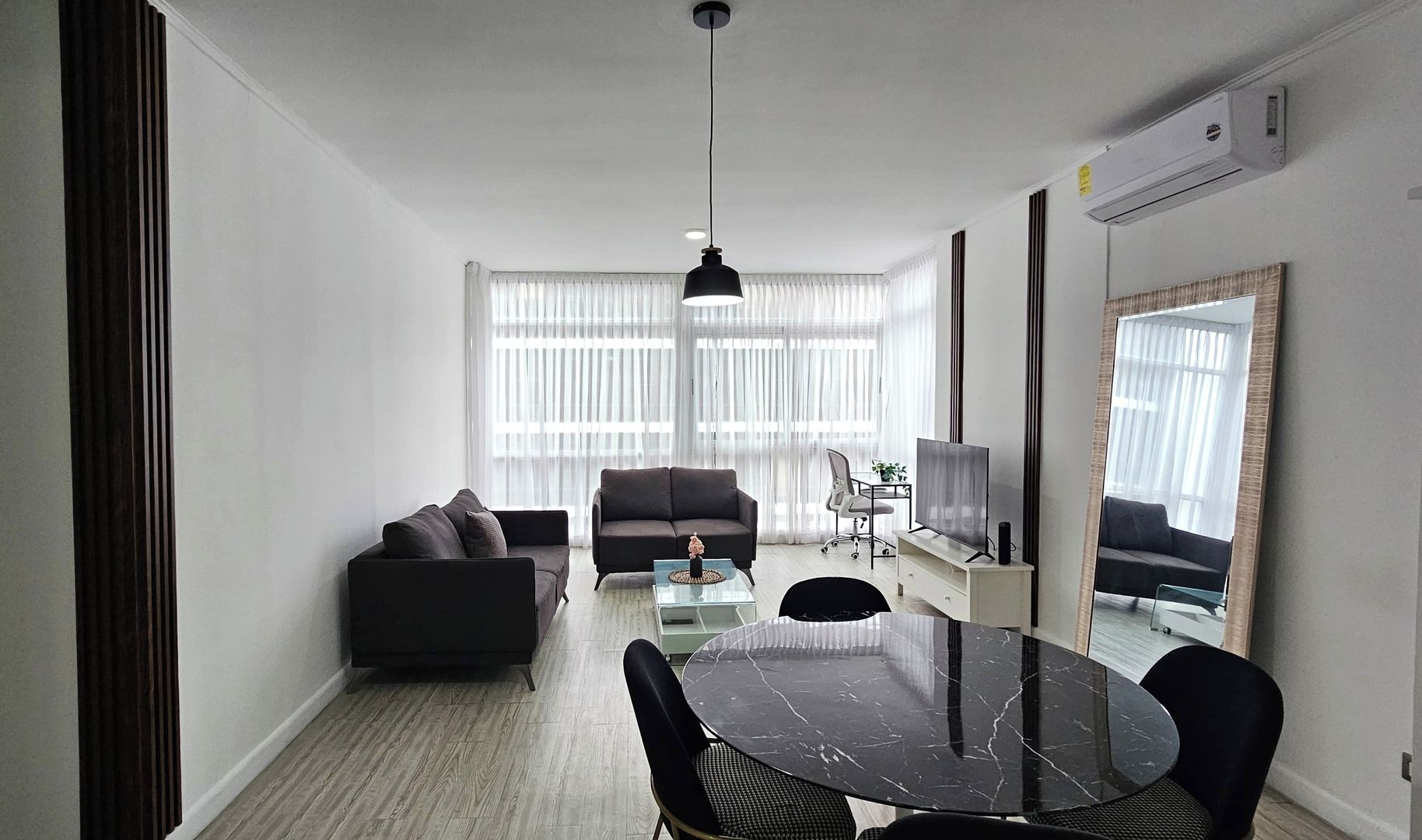 A modern living room with a sofa, a dining table with chairs, a TV, and a large window with sheer curtains.