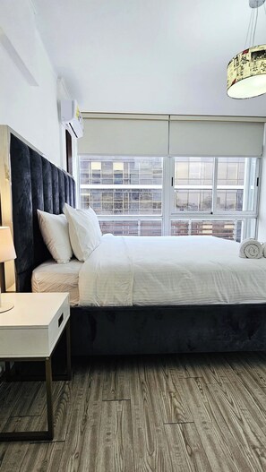 A bedroom with a bed, bedside table, lamp, and a window with a view of buildings.