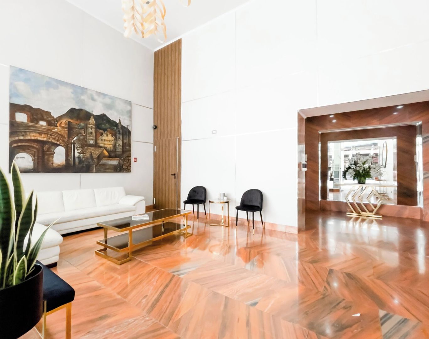 Un vestíbulo de hotel moderno con un sofá blanco, dos sillas negras, una mesa de centro de vidrio y un cuadro grande de un edificio histórico.