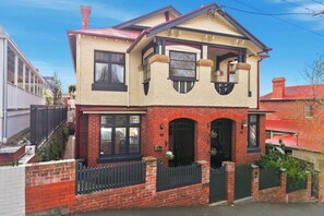A two-story house with a red brick facade, a balcony, and a black metal fence. - Ethelea Retreat – 2 Bedroom – Walk to Restaurant Precinct (North Hobart)