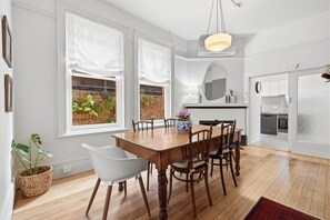 A dining area with a wooden table and chairs, a hanging light fixture, and a view of a brick wall and greenery through the windows. - Ethelea Retreat – 2 Bedroom – Walk to Restaurant Precinct (North Hobart)