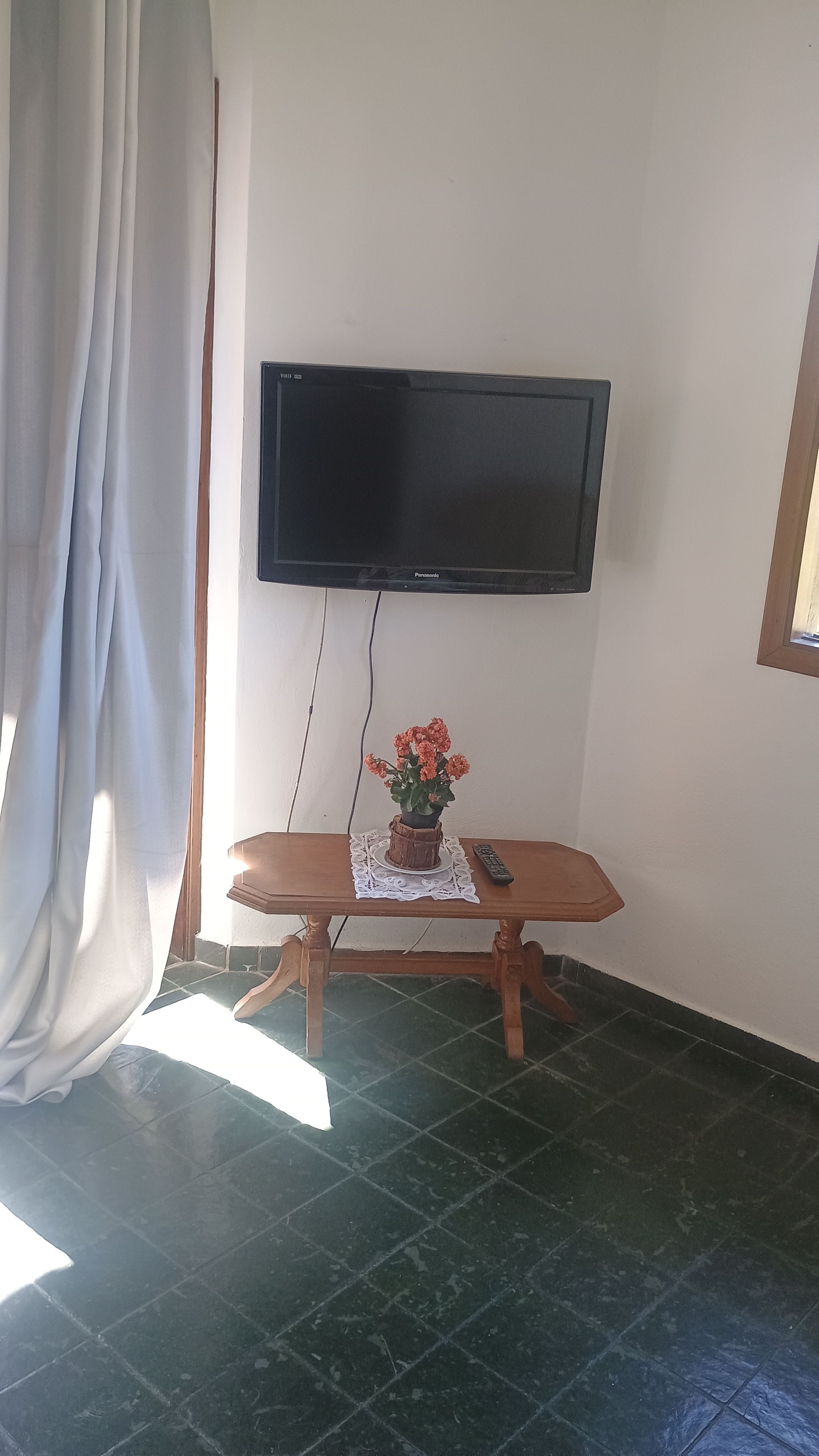 Quarto com televisão de tela plana na parede, mesa de madeira com toalha de renda e planta em vaso.