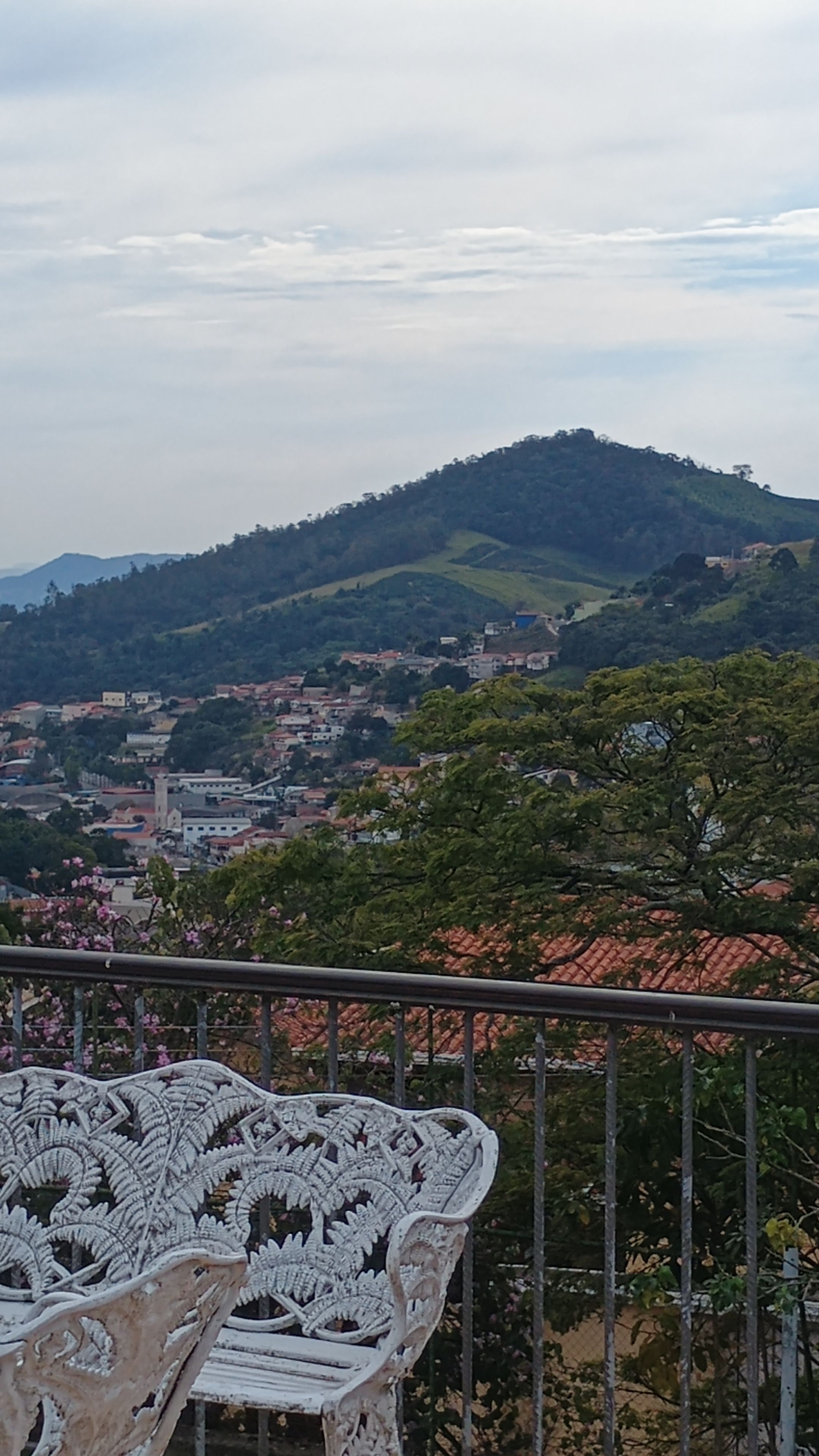 A balcony with a white ornate chair, overlooking a town with buildings and hills in the distance.