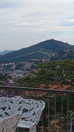 Un balcón con una silla blanca ornamentada, con vistas a un pueblo con edificios y colinas al fondo.