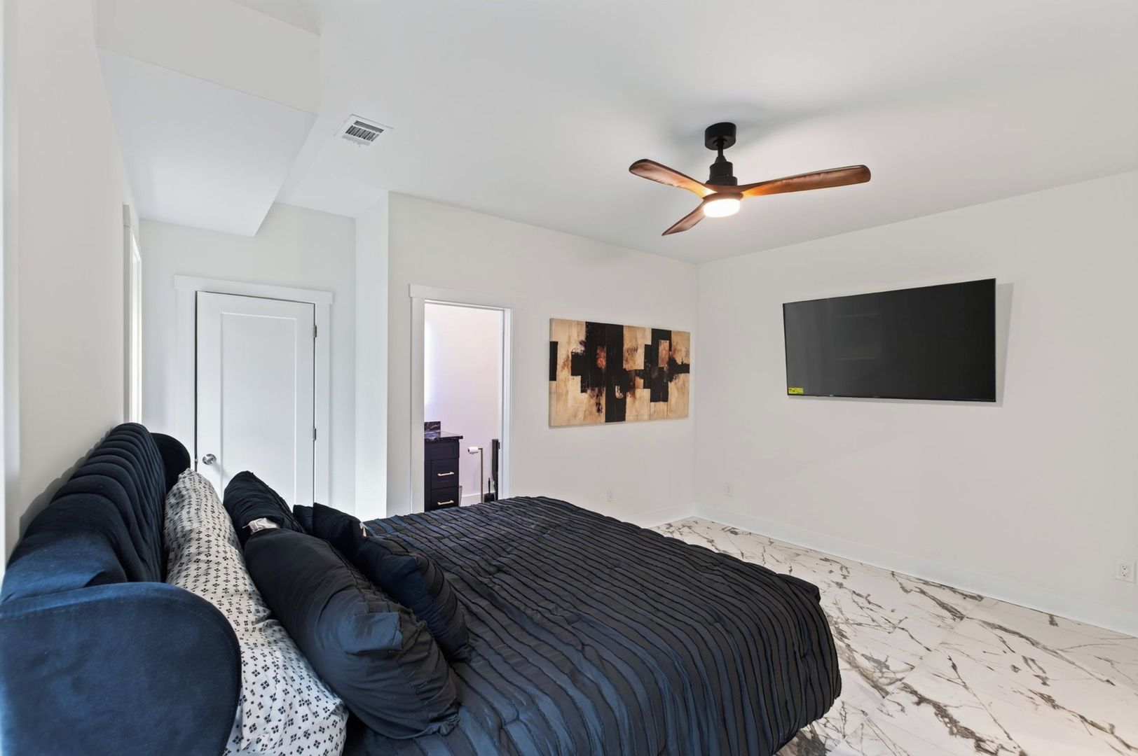 A bedroom with a bed, a ceiling fan, a wall-mounted TV, and a door leading to another room.