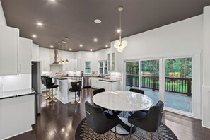 A modern kitchen with a central island, dining area with a round table, and a view of a deck.