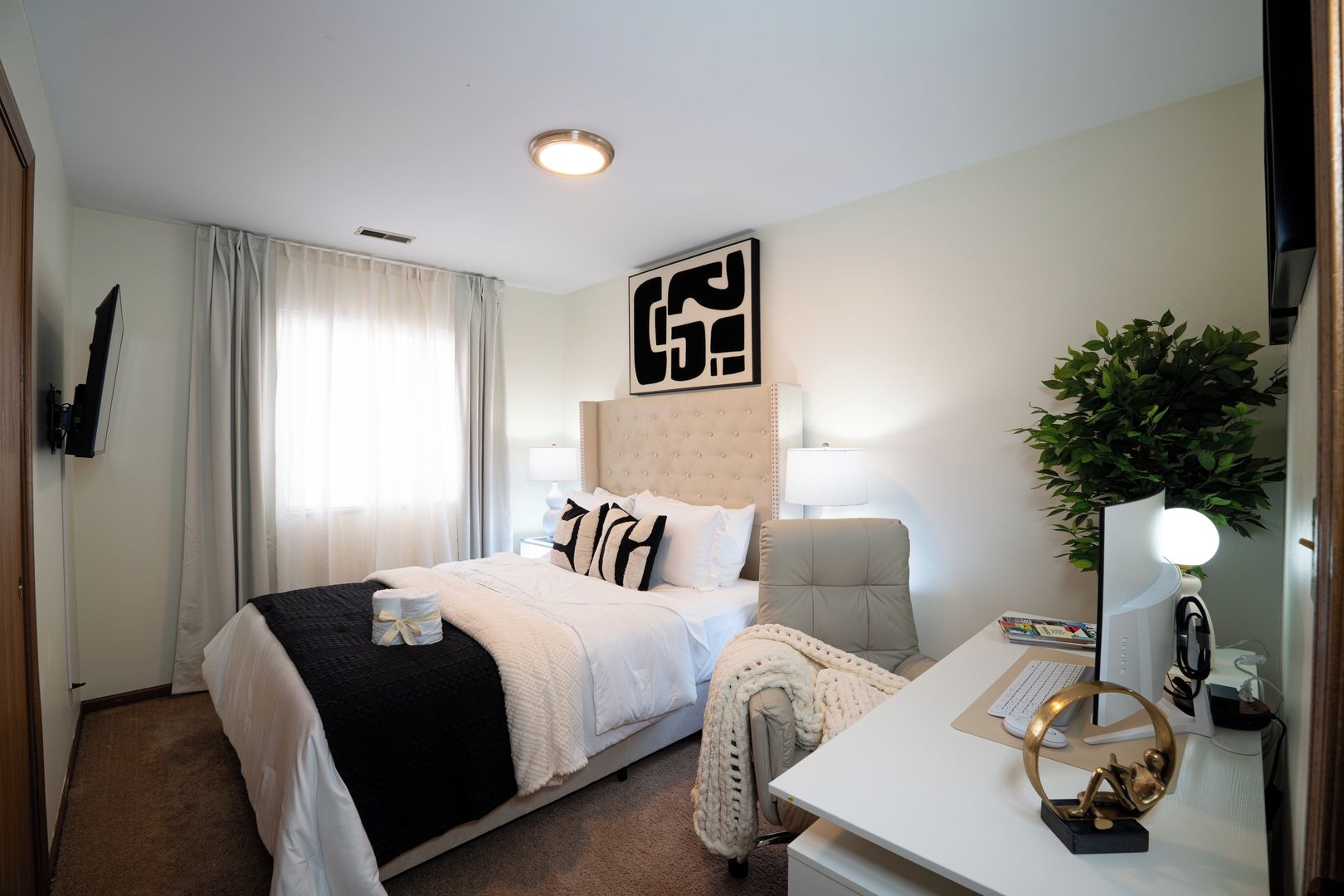 A hotel room with a bed, a desk, a chair, a lamp, a plant, and a wall-mounted artwork.