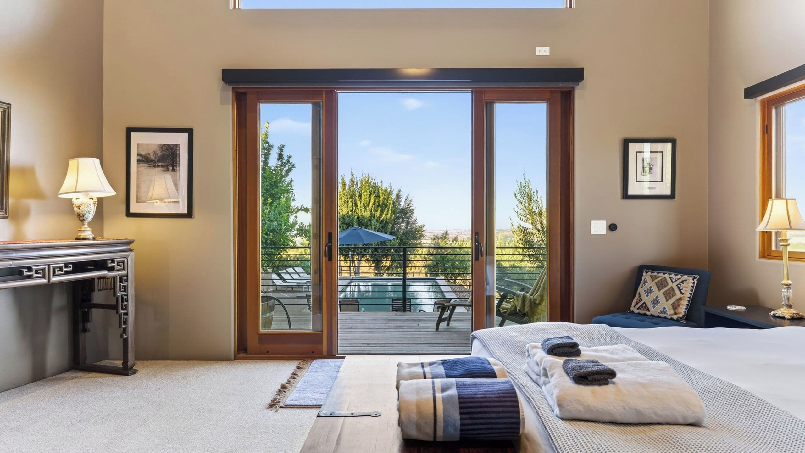 A bedroom with a large sliding glass door leading to an outdoor deck, a bed with a view, and a desk with a lamp.