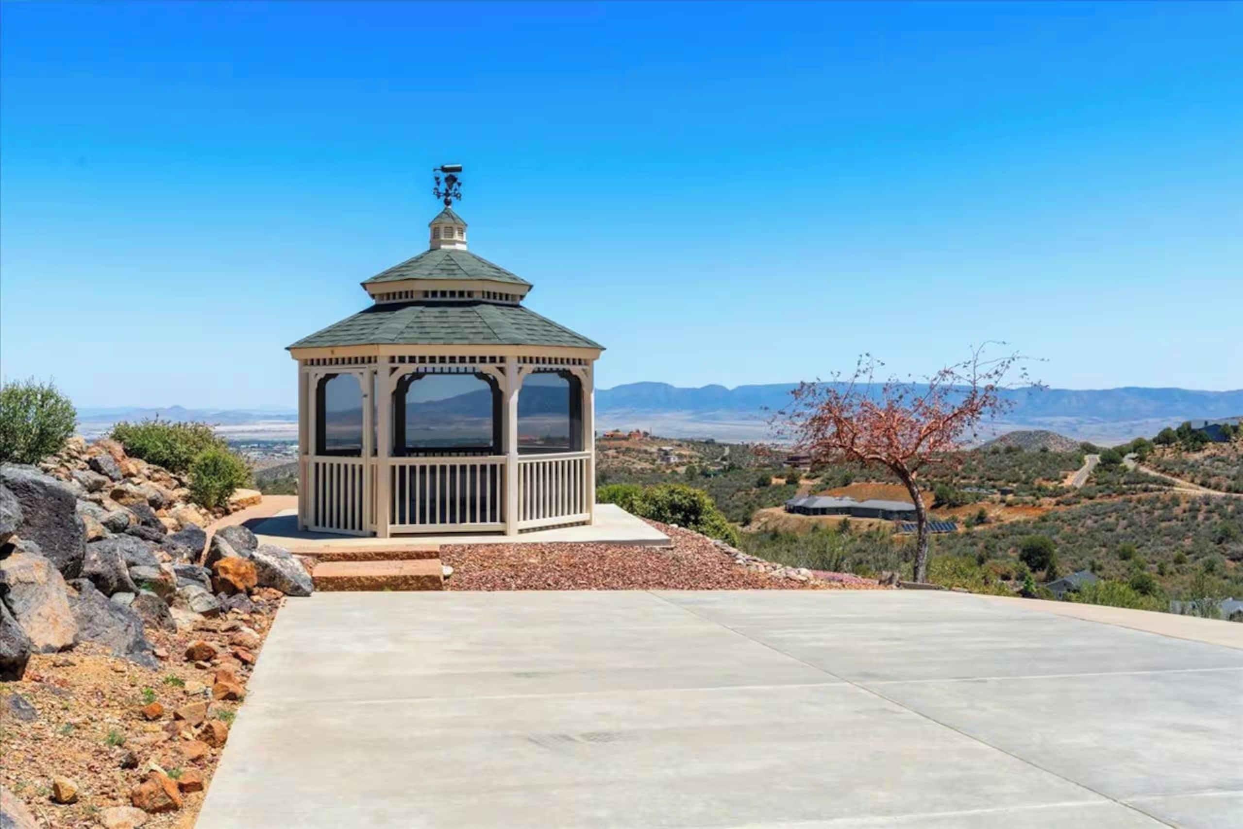 A gazebo with a dome roof and a small tower, surrounded by a paved area and a natural landscape with hills and sparse vegetation.