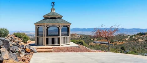 A gazebo with a dome roof and a small tower, surrounded by a paved area and a natural landscape with hills and sparse vegetation.