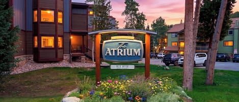 A hotel sign with 'The Atrium' prominently displayed, surrounded by a landscaped garden with various flowers and a parking area with several cars.