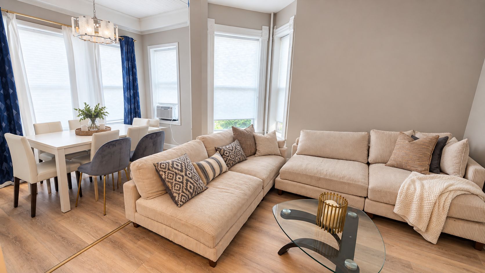 Ein modernes Wohnzimmer mit einem beigefarbenen Sofa, einem Glastisch und einem Essbereich mit weißem Tisch und Stühlen.