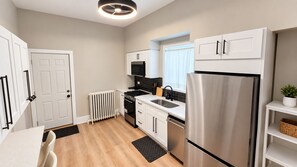 A modern kitchen with white cabinetry, a stainless steel refrigerator, a black stove, and a window with a white curtain. - Duplex - Both Units - 2 Kitchens - Privacy - Fenced Yard (Oak Park)