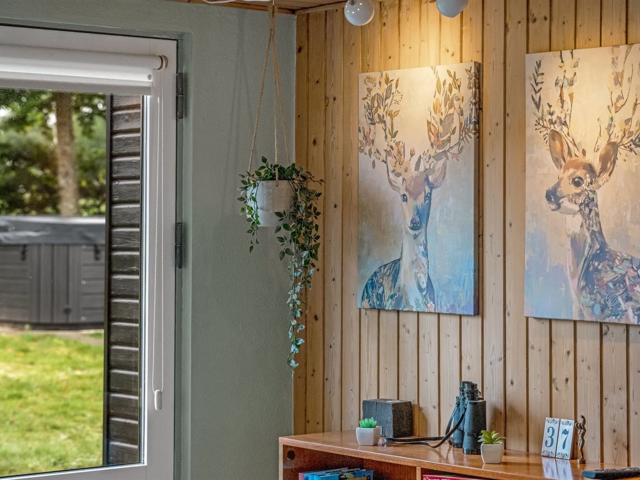 A room with wooden panel walls, a window with blinds, a hanging plant, two deer paintings, a telescope, and a small table with a plant and a number sign.