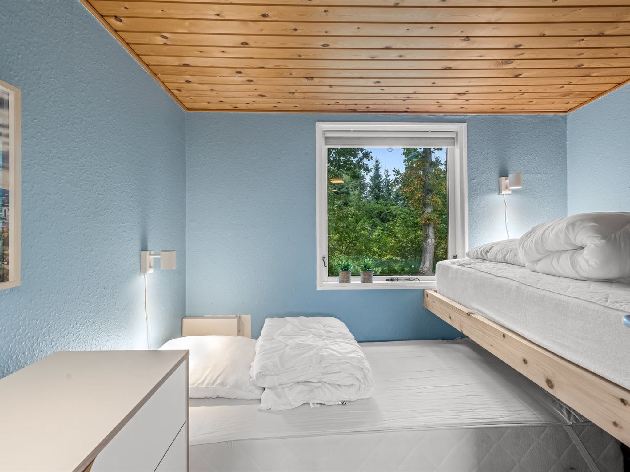 A bedroom with a bunk bed, a bedside table, a desk, and a window with a view of trees.