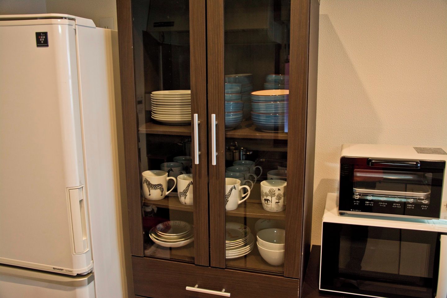 A kitchen with a refrigerator, a microwave, and a glass-fronted cabinet with dishes and mugs.