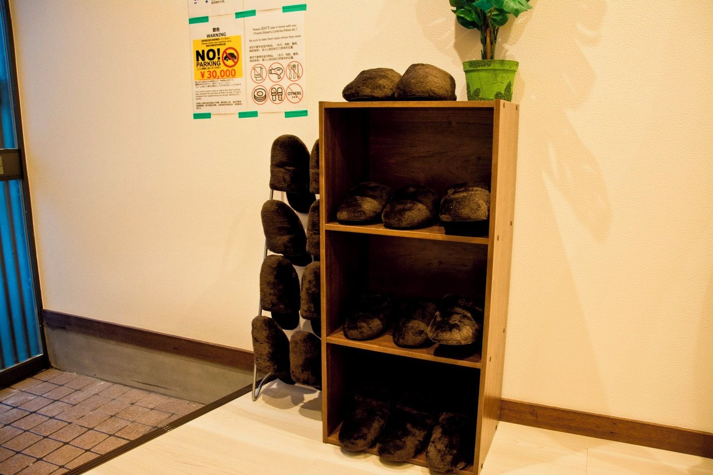 A wooden shoe rack with multiple shelves against a wall, next to a no parking sign and a potted plant.