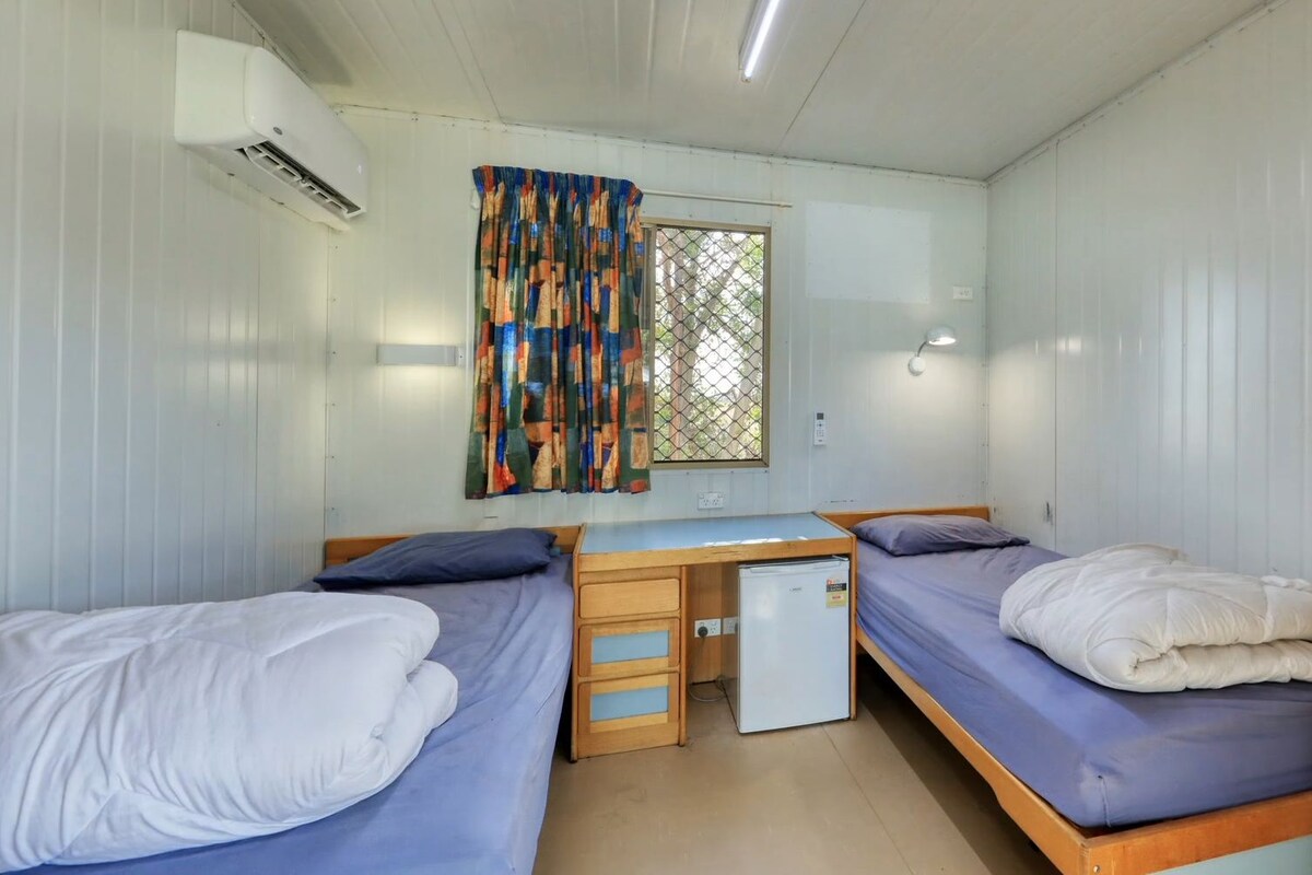 A compact room with two beds, a small desk, a refrigerator, and a window with a colorful curtain.