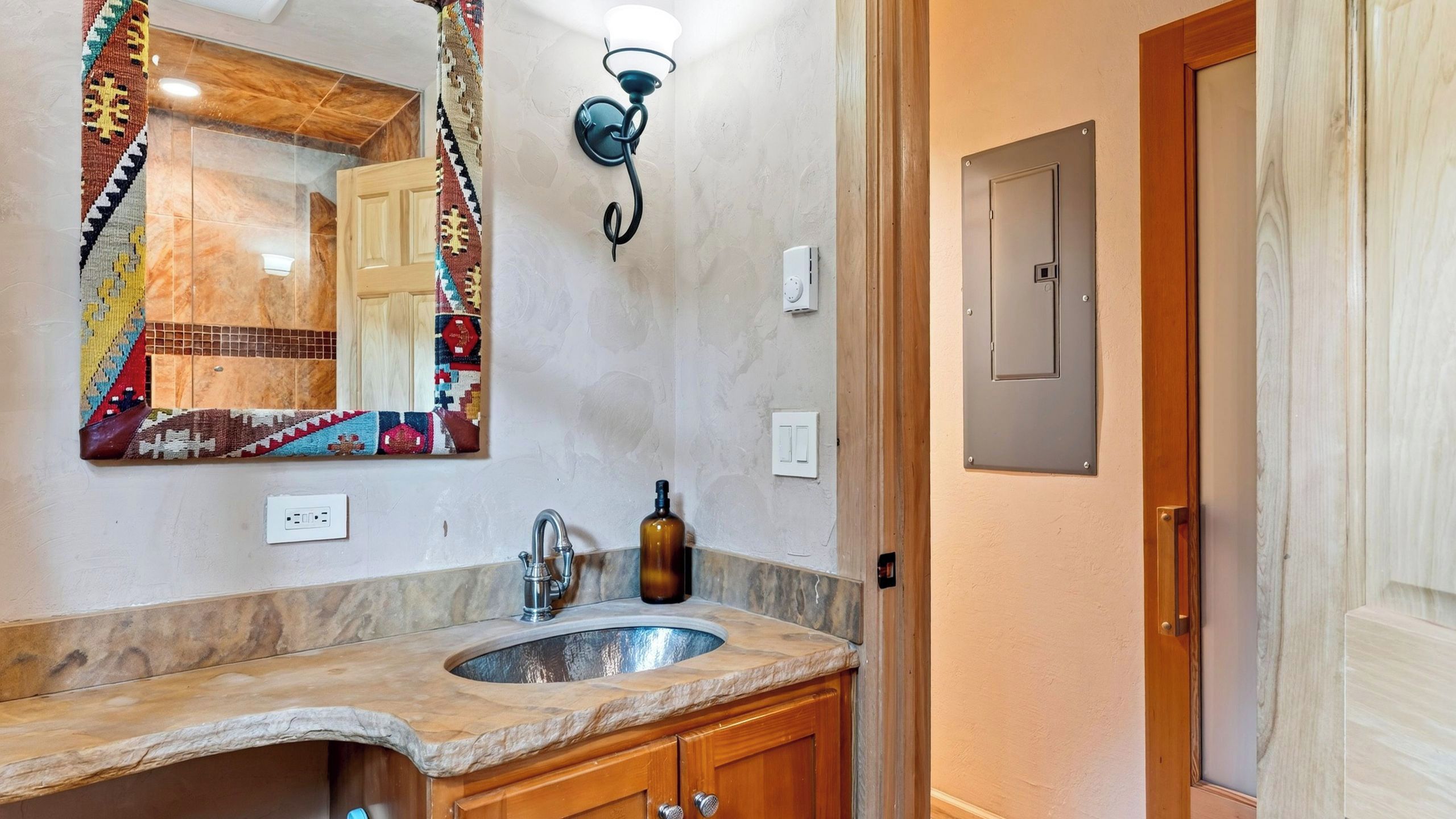 A bathroom with a marble sink, wooden cabinets, a mirror with a colorful border, a wall-mounted light fixture, and a door with a panel.