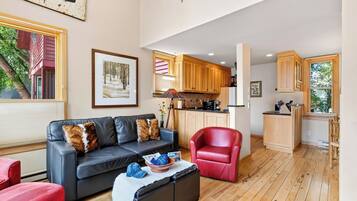 Una sala moderna con un sofá de cuero, un sillón rojo y una zona de comedor con armarios de madera y una cocina al fondo.
